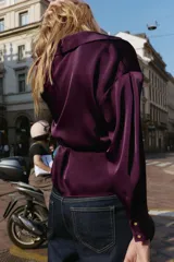 Camisa satinada de color violeta oscuro, con cuello solapa, escote en pico y manga larga. Presenta un detalle de cinturón fino ajustable a tono en la cintura y cierre frontal con botones.