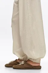 Zapatilla estilo mula de color beige, confeccionada en una combinación de algodón y serraje vacuno, con suela de caucho con tachuelas texturizadas y costuras decorativas visibles en la parte superior.