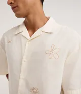 Camisa masculina boxy de manga corta en tricolina color marfil, con cuello cubano, cierre frontal con botones y sutiles bordados veraniegos en tono tostado.