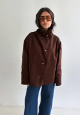 Campera marrón de 100% algodón, con cuello alto estructurado y cierre frontal cruzado con botones metálicos a presión. Presenta diseño holgado y de corte recto.