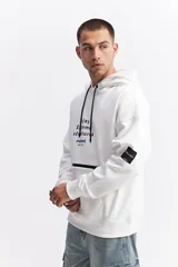 Buzo tipo canguro oversize de color blanco, con capucha, bolsillo frontal y estampado gráfico en el pecho. Presenta un parche rectangular negro en la manga y cordones ajustables en la capucha.