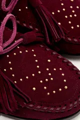 Mocasín de gamuza color bordó con detalle de flecos, cordones y tachas metálicas en la parte superior.