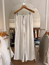Pantalón palazzo blanco de lino, con cintura elástica fruncida y botón frontal decorativo. Presenta un corte amplio y relajado de pierna ancha.