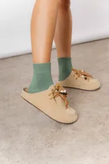 Zueco de cuero gamuzado color tostado, con cordones decorativos adornados con cuentas de madera y piezas envueltas en cuero. Diseño tipo mocasín con costuras visibles en la parte superior y abierto en la parte trasera.