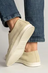 Zapatillas de gamuza color arena, diseño sin cordones estilo slip-on, con costuras decorativas en el empeine y suela de goma blanca.