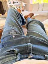 Pantalón de jean azul con corte holgado y detalle de tres cruces bordadas en el muslo derecho. Presenta costuras reforzadas y cierre frontal con botón metálico.