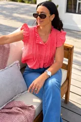 Blusa de diseño cuadrillé en color rojo y blanco, sin mangas, con volados en el cuello mao y en los hombros. Cuenta con alforzas verticales en el frente y cierre frontal de botones.