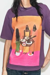 Remera oversize de algodón con efecto lavado en color violeta. Presenta cuello redondo, manga corta y una estampa frontal colorida con una ilustración artística de Manuja Waldia.