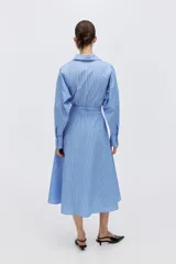 Vestido midi camisero de rayas azules y blancas, con cuello clásico, mangas largas, cierre frontal de botones y lazo ajustable en la cintura.
