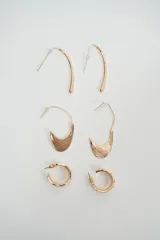 Set de tres pares de caravanas doradas de diferentes diseños: un par de aros alargados y curvos, un par de aros con dije en forma de media luna texturizada y un par de aros tipo argolla pequeña.