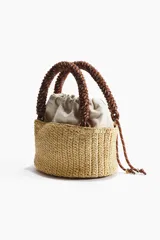 Cartera tipo tote en paja sintética trenzada, con estructura cilíndrica. Cuenta con un bolso interior de lona color beige con cordón ajustable, asa superior trenzada de cuerda marrón y detalles en ecuero.