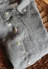 Camisa gris de lino natural con doble cuello, cartera escondida y tablas frontales. Presenta delicados bordados de flores en cuello y frente.