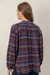 Camisa de corte holgado con estampado de cuadros escoceses en tonos azul, rojo y marrón. Presenta cuello mao, cierre frontal con botones y un detalle de volado vertical a lo largo de la cartera. Mangas largas con puños abotonados.