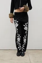Pantalón de denim negro de corte recto con estampado floral blanco en ambas piernas. Cuenta con cierre frontal y bolsillos laterales.