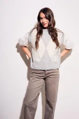 Chaleco de tejido de punto con diseño de rombos en tonos gris claro y blanco. Presenta mangas abullonadas de tela blanca con detalles de encaje y cuello redondo.