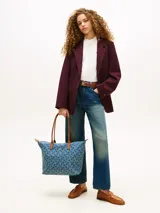 Cartera tote Tommy Hilfiger confeccionada en tejido denim azul con estampado geométrico de monogramas. Cuenta con asas superiores de cuero sintético marrón, cierre superior de cremallera y herrajes metálicos dorados con el monograma de la marca en el frente.