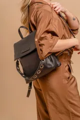 Mochila tipo cartera con tapa, confeccionada en símil cuero color suela. Cuenta con doble compartimento, división interna, cierre de seguridad y bolsillo trasero con cierre. Incluye una correa frontal decorativa con tachas metálicas.