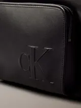 Mochila negra de diseño moderno con acabado tipo piel sintética. Presenta un asa de mano superior, correas ajustables para el hombro, compartimento principal con cremallera y un bolsillo exterior frontal con el monograma 'CK' grabado en relieve.