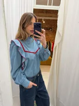 Blusa de jean fino con cuello alto, mangas largas con volumen y volados decorativos en los hombros. Presenta detalles de ribetes en terciopelo azul oscuro en el frente y los volados.