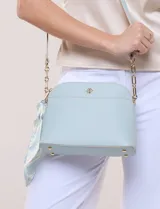 Cartera bandolera estructurada de eco-cuero verde claro, con silueta curva, bolsillo frontal y detalle de pañuelo estampado atado en el lateral. Posee correa larga con detalles de cadena dorada.