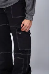 Pantalón tipo carpintero de corte relaxed fit y tiro medio, confeccionado en denim negro con costuras a contracolor en blanco. Presenta múltiples bolsillos cargo laterales, un bolsillo con cierre en la parte inferior y presillas utilitarias.