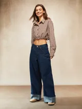 Camisa de manga larga, cuello camisero y botones frontales, con estampado a cuadros marrones y blancos. El ruedo inferior es ajustable con cordones laterales, creando un efecto *cropped* y fruncido.