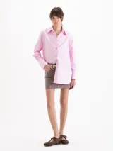 Camisa holgada de poplin blanco, con cuello clásico, mangas largas con puños anchos y cierre frontal con botones. Presenta detalles de apliques metálicos en las puntas del cuello y sutil bordado en el delantero.