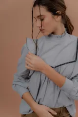 Blusa de jeans fino color celeste con detalles de terciopelo negro en contraste. Presenta cuello alto con volados, mangas largas abullonadas con puños ajustados y volados decorativos en los hombros y el frente.