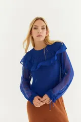 Blusa azul de crepe con transparencias, cuello redondo con volados y mangas largas con puños ajustados.