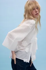 Camisa de tejido fluido color blanco, con cuello solapa, cierre frontal con botones y mangas amplias por debajo del codo. Presenta aplicaciones de flores en relieve a tono distribuidas por toda la prenda.