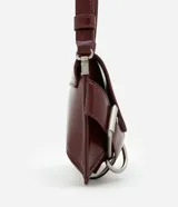 Cartera baguette Satinato pequeña de color negro, confeccionada en poliuretano. Presenta una solapa frontal con cierre de botón a presión, correa ajustable para el hombro y detalles decorativos de hebillas con argollas metálicas plateadas en los laterales.