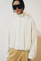 Chaqueta de nylon color crudo, con cuello alto, mangas largas, bolsillos delanteros, bajo abullonado con elástico y cierre frontal con botones forrados.