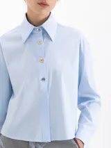 Camisa de corte recto y holgado, color celeste, con cuello clásico, cierre frontal con botones y mangas largas con puños abotonados.