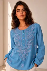 Blusa blanca de manga larga con detalles bordados en tono marrón en el pecho y las mangas, cuello redondo y cierre frontal con pequeños botones.