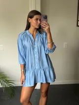 Vestido corto azul claro de manga larga, con cuello mao y cierre de botones frontal. Presenta detalles de encaje o bordado en las costuras verticales del frente y en la zona de los hombros. La manga se ajusta con lazo y el ruedo tiene un volante fruncido.