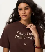 Remera de algodón color marrón oscuro, con cuello redondo y manga corta. Presenta un estampado frontal en blanco con el texto "Sunday Club Palm Paradise", una palmera y una frase motivacional.