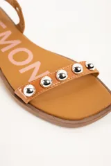 Sandalias planas de cuero color suela, con tira en la puntera y tira en el empeine decoradas con tachas semiesféricas plateadas, y tira ajustable con hebilla en el tobillo.