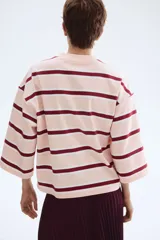Remera de manga tres cuartos con cuello redondo, hombros caídos y corte holgado. Presenta un patrón de rayas horizontales en tonos rosa pálido, blanco y granate.