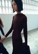 Vestido midi de corte evasé en color marrón oscuro, confeccionado en tejido elástico. Presenta cuello asimétrico, manga larga y diseño sin cierre.