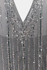 Vestido corto gris, estilo flapper o años 20, con cuello halter y escote pronunciado en pico. Confeccionado en tul con apliques verticales de lentejuelas y brillos plateados. Presenta un ruedo con volado.