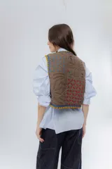 Chaleco de algodón color marrón con bordados decorativos en tonos azul, amarillo y fucsia. Presenta un diseño corto con cierre frontal mediante tiras para atar y detalles de borlas.