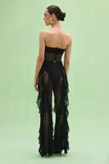 Mono strapless negro confeccionado en encaje con diseño calado. Presenta un corpiño con detalle de frunce y hebilla dorada en el centro, cintura marcada con cinturón fino y hebilla dorada, y piernas acampanadas con volados laterales de encaje.