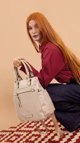 Cartera mochila de diseño versátil en color beige, confeccionada en tela impermeable. Cuenta con doble asa superior para llevar en la mano, correas ajustables para usar como mochila, bolsillo frontal con cierre y bolsillos laterales abiertos.