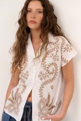 Camisa de manga corta color beige con cuello camisero, cierre frontal con botones y bordado marrón de motivos florales y vegetales.