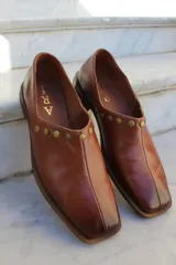 Zapato tipo mocasín de cuero vacuno color marrón, con punta cuadrada y detalle de tachas metálicas planas en el borde superior. Presenta una costura central vertical y un taco geométrico.