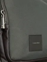 Bolso tipo reportero o bandolera de Calvin Klein, color verde oliva oscuro con detalles en marrón oscuro. Presenta un diseño funcional con un bolsillo exterior frontal con cierre y un parche de cuero sintético con el logo de la marca. Incluye correa ajustable de tela.