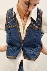 Chaleco de denim azul con bordados metalizados y pedrería en la parte superior delantera, escote en V y cierre frontal con botones metálicos.