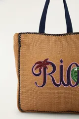Bolso tote de mano y hombro, color marrón con textura tejida de efecto natural, asas dobles azules y bordado frontal con la palabra 'Rio' decorada con una palmera y un tucán.