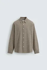 Camisa de corte relajado confeccionada en tejido de algodón con textura irregular y estampado de cuadros pequeños en tonos tierra. Presenta cuello solapa, cierre frontal de botones y mangas largas con puños abotonados.