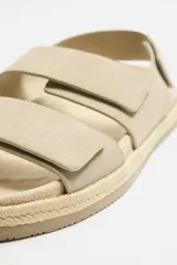 Sandalia color beige de piel con acabado serraje. Cuenta con dos tiras anchas en el empeine y tira en talón ajustables mediante cierres adherentes. Plantilla con horma anatómica y piso forrado de yute.